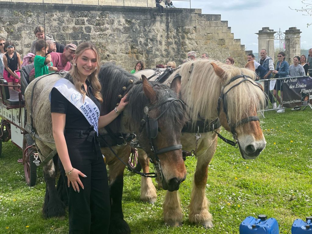 Une jeune femme vêtue d'une tenue sombre avec une écharpe de Miss Picar'naval posant avec deux chevaux dans un parc, devant un public rassemblé.